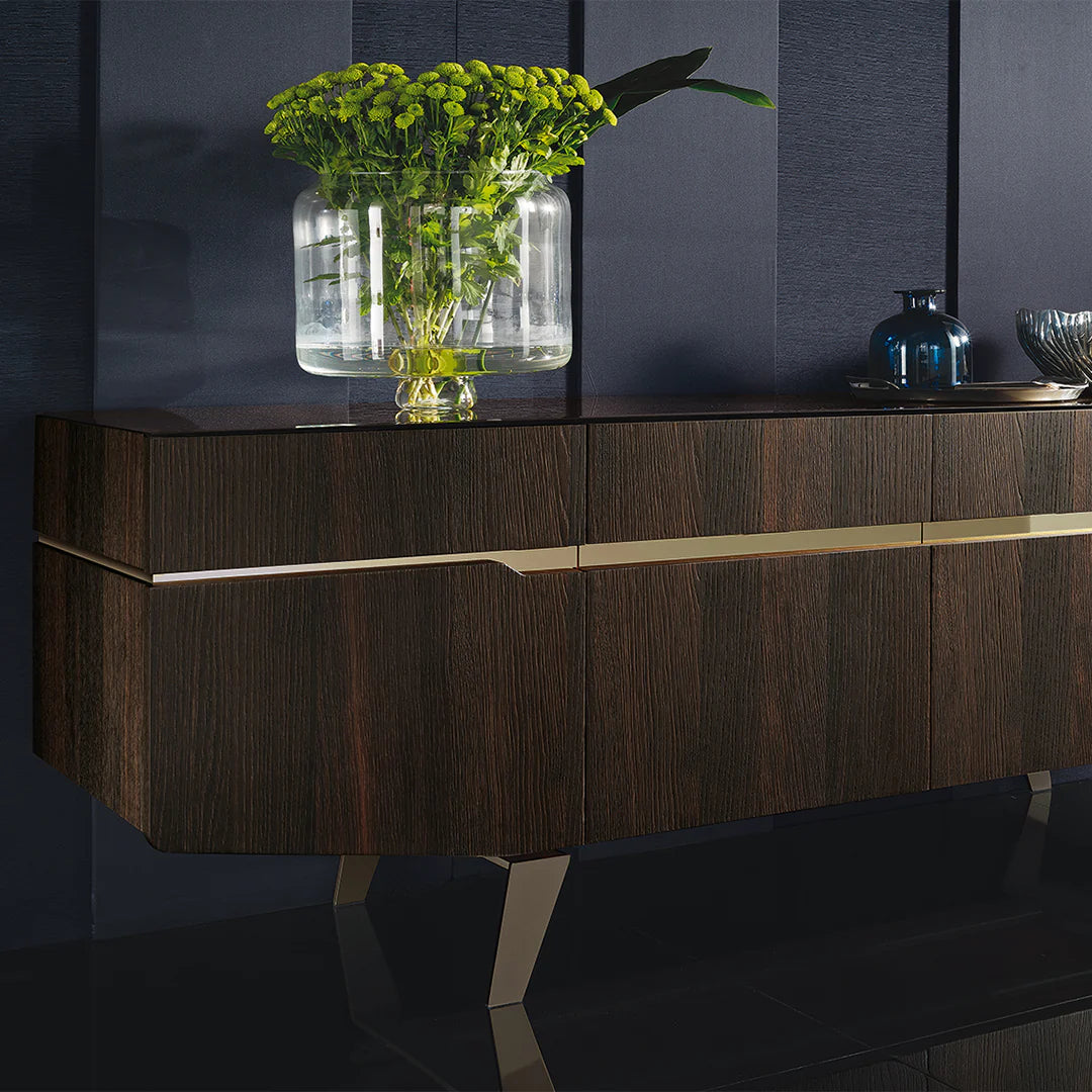 A luxury dark wood sideboard with gold metal accents and legs, styled with a glass vase of green flowers and decorative blue