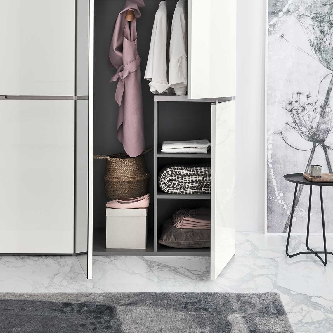 A white minimalist wardrobe filled with folded clothes, hanging shirts, and woven baskets is shown in a bright, modern room w